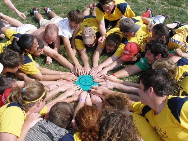 A coed frisbee team lays on the ground in a circle around a frisbee.