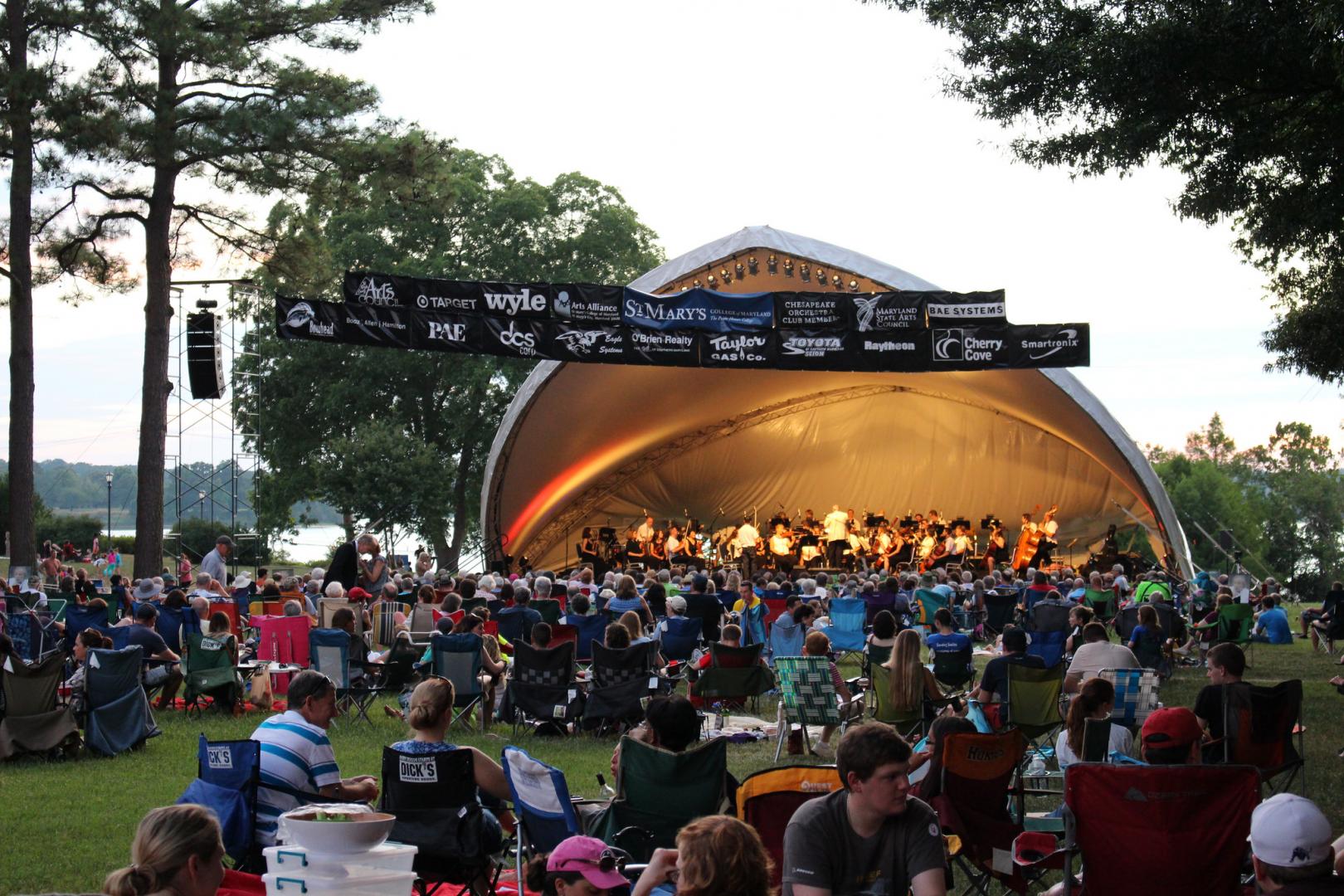 A crowd of concert goers watch the orchestra under a tent in front of the St. Mary's River