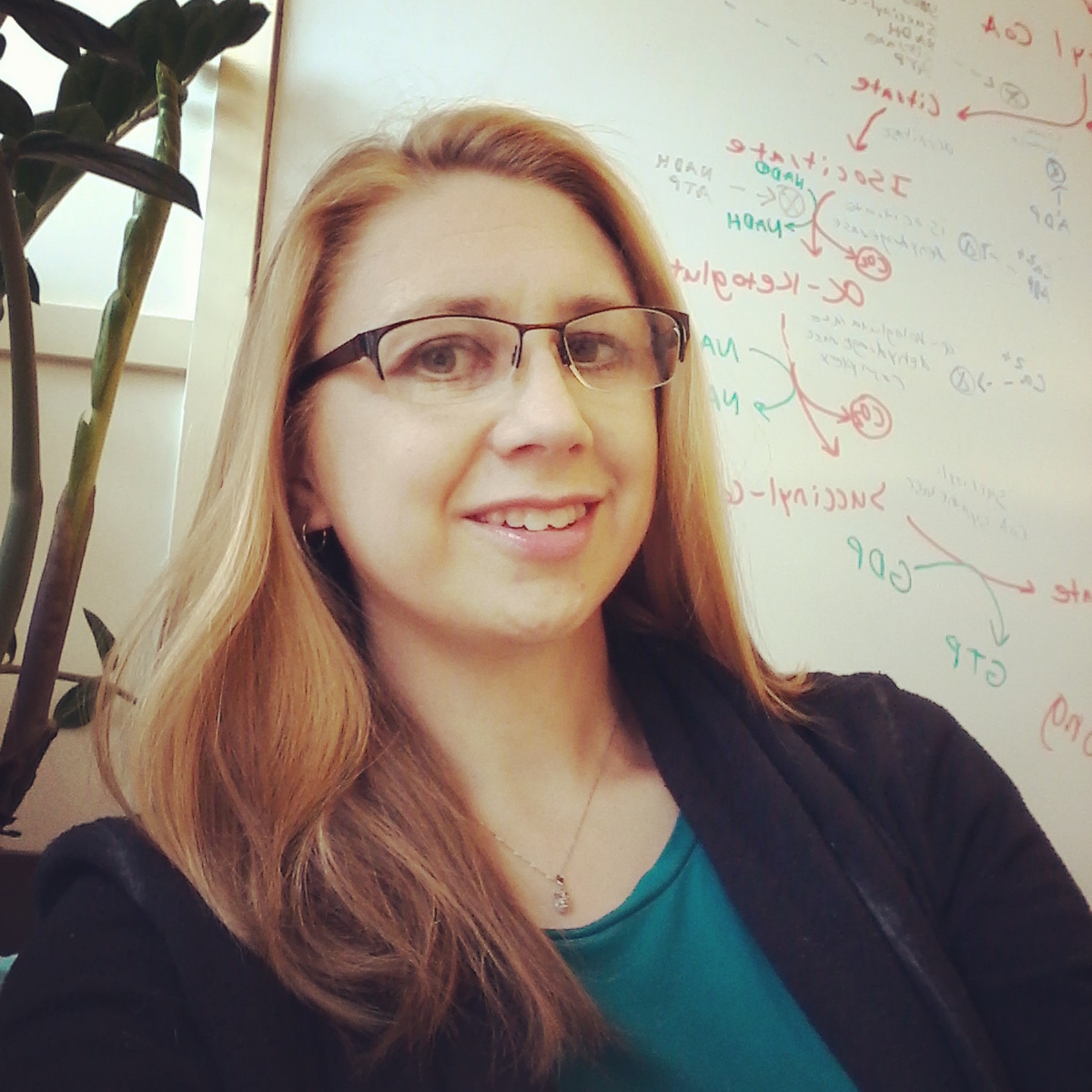 A white woman with glasses and long hair sits in front of a white board with a biochemical cycle sketched on it.