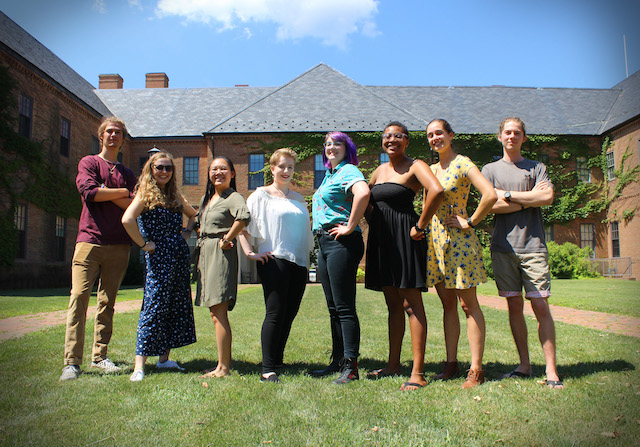 8 confident looking diverse students stand on the lawn in front of an academic building