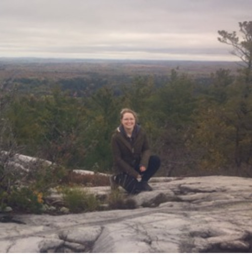Morgan Siewert kneeling on a stone cliff overlooking an evergreen forest