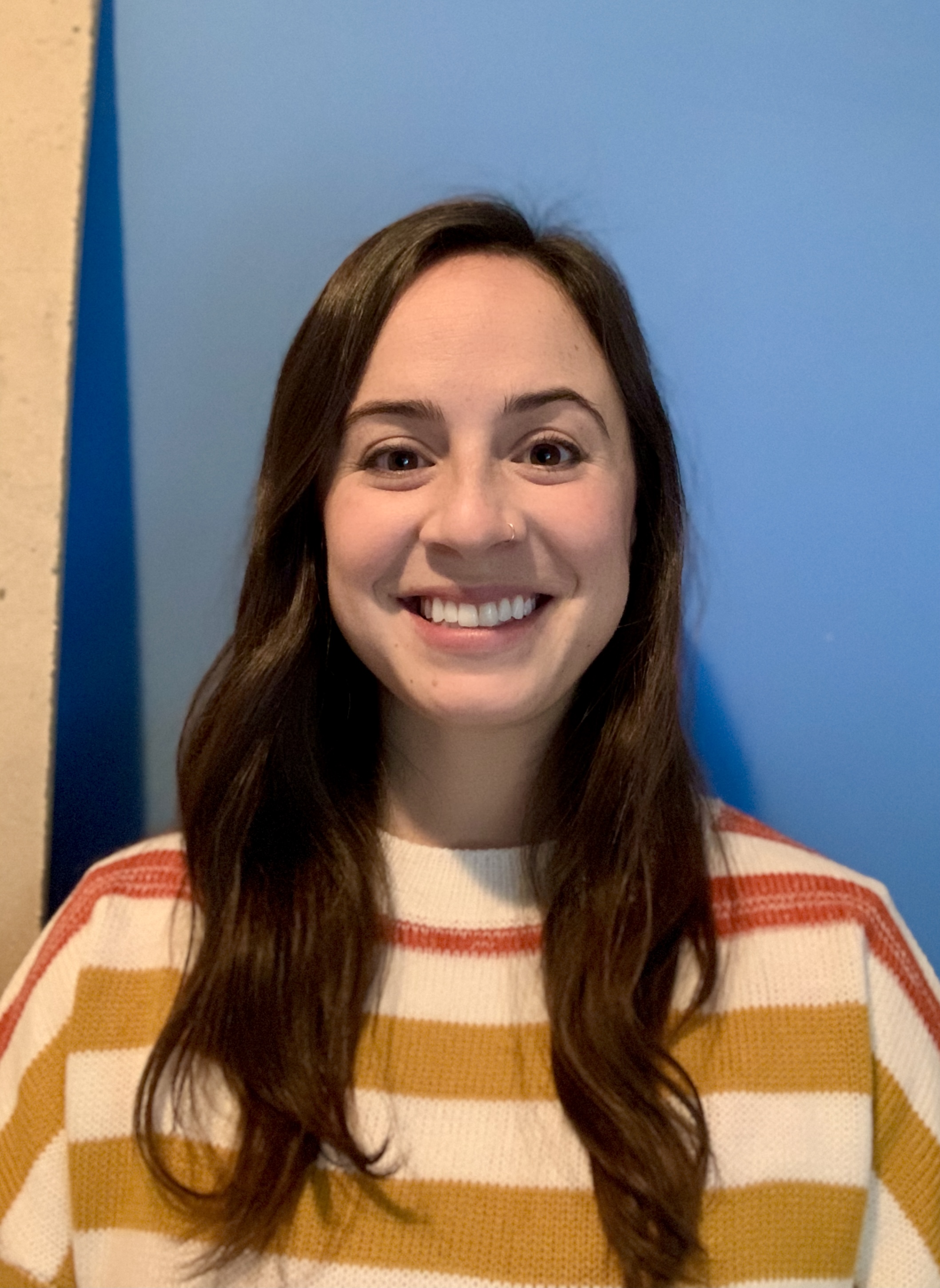 Rachel Heiss is smiling at the camera, wearing an orange, yellow and white striped sweater and standing in front of a blue wall.