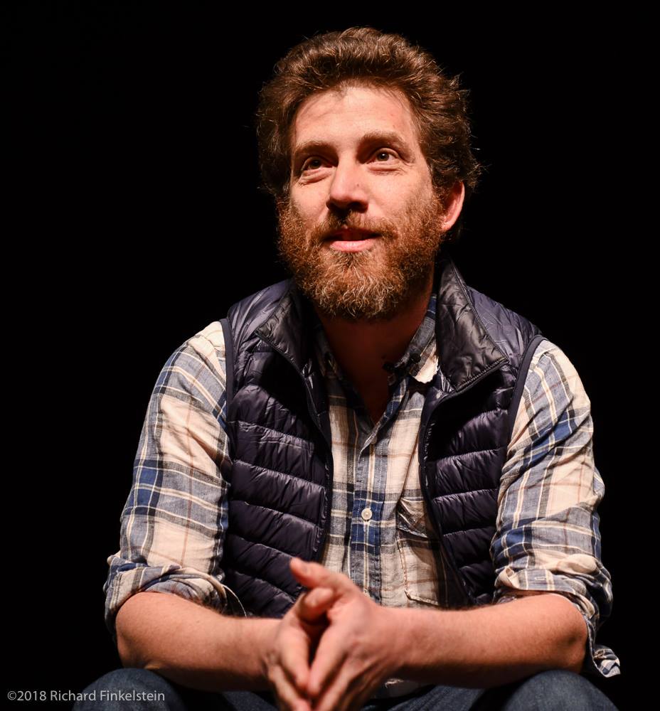 Julian Boal, a bearded man with red hair, is pictured against a black background wearing a flannel shirt and vest jacket. He is seated with his hands folded looking straight ahead.
