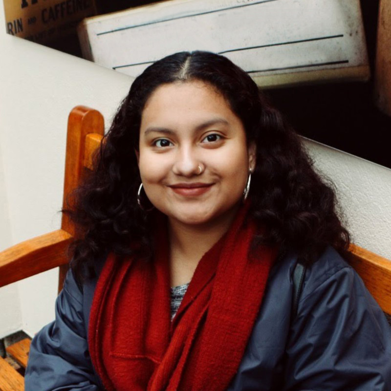 person with wavy brown hair wearing orange scarf and dark navy jacket sitting on bench.