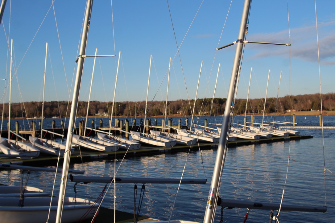 Angled photo of sailboats stationary on the floating docks, without sails. The sky is bright blue and the water is calm. 