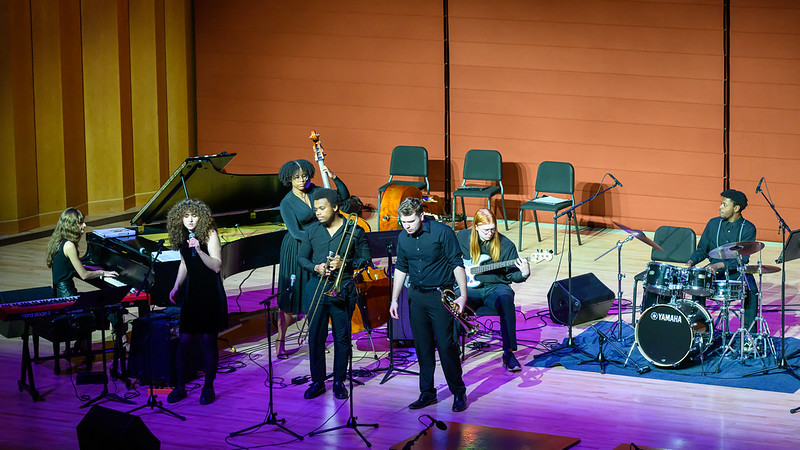 Students perform on stage on piano, brass, bass, drums and signing