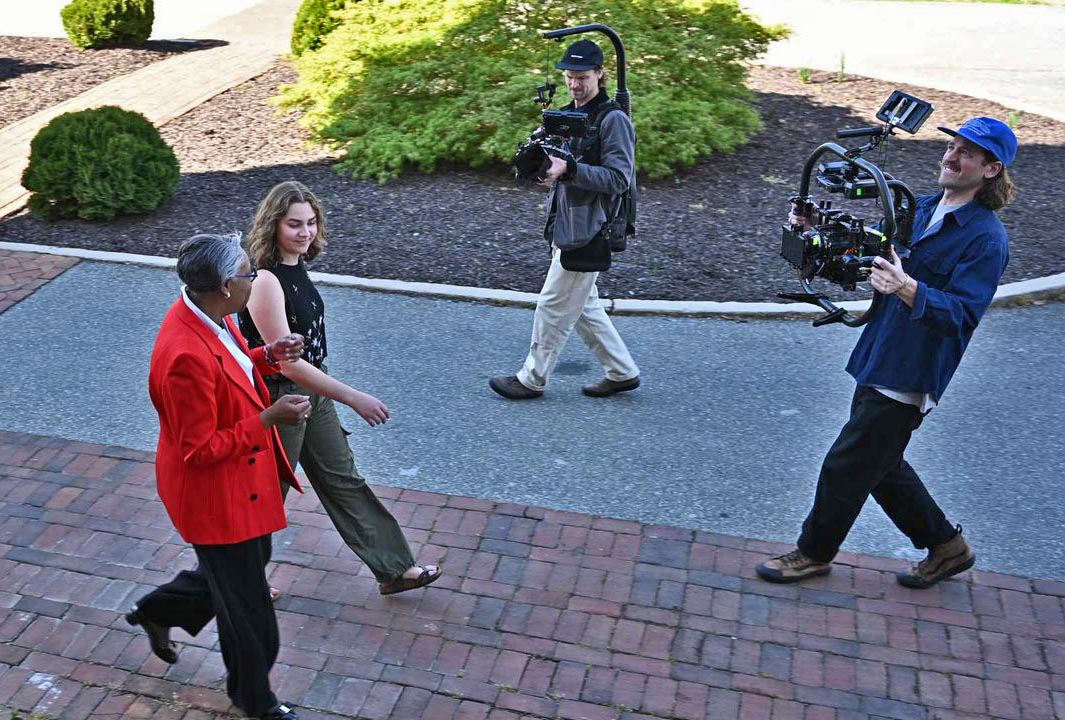 Two people walk in front of two video cameramen