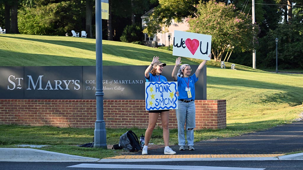 Students holding welcome signs wave to cars at campus entrance