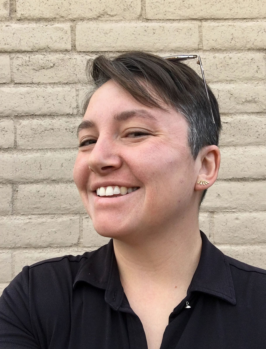 A woman with short brown hair, wearing a black button-down shirt, stands in front of a brick wall. She is smiling, wearing glasses on top of her head. 