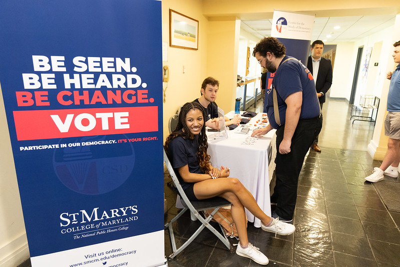 Students sit at a table outside the great room next to a banner that reads: "Be Seen. Be Heard. Be Change. Vote."