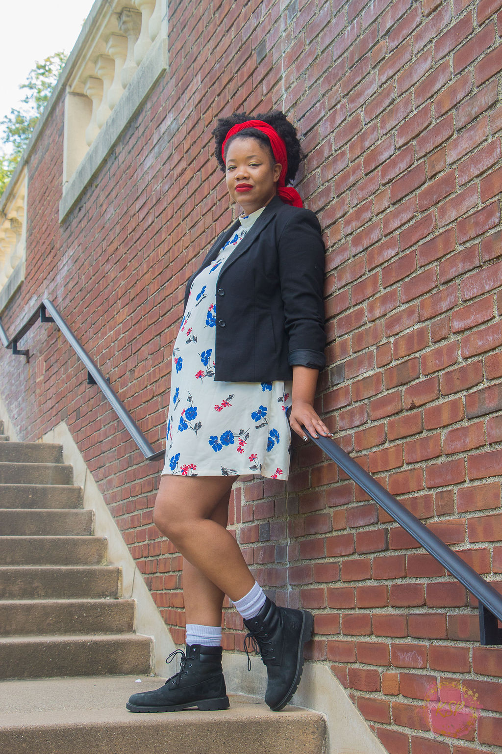 Black woman with red head wrap, natural black hair, wearing a black blazer, a white dress with red and blue flowers, and black boots