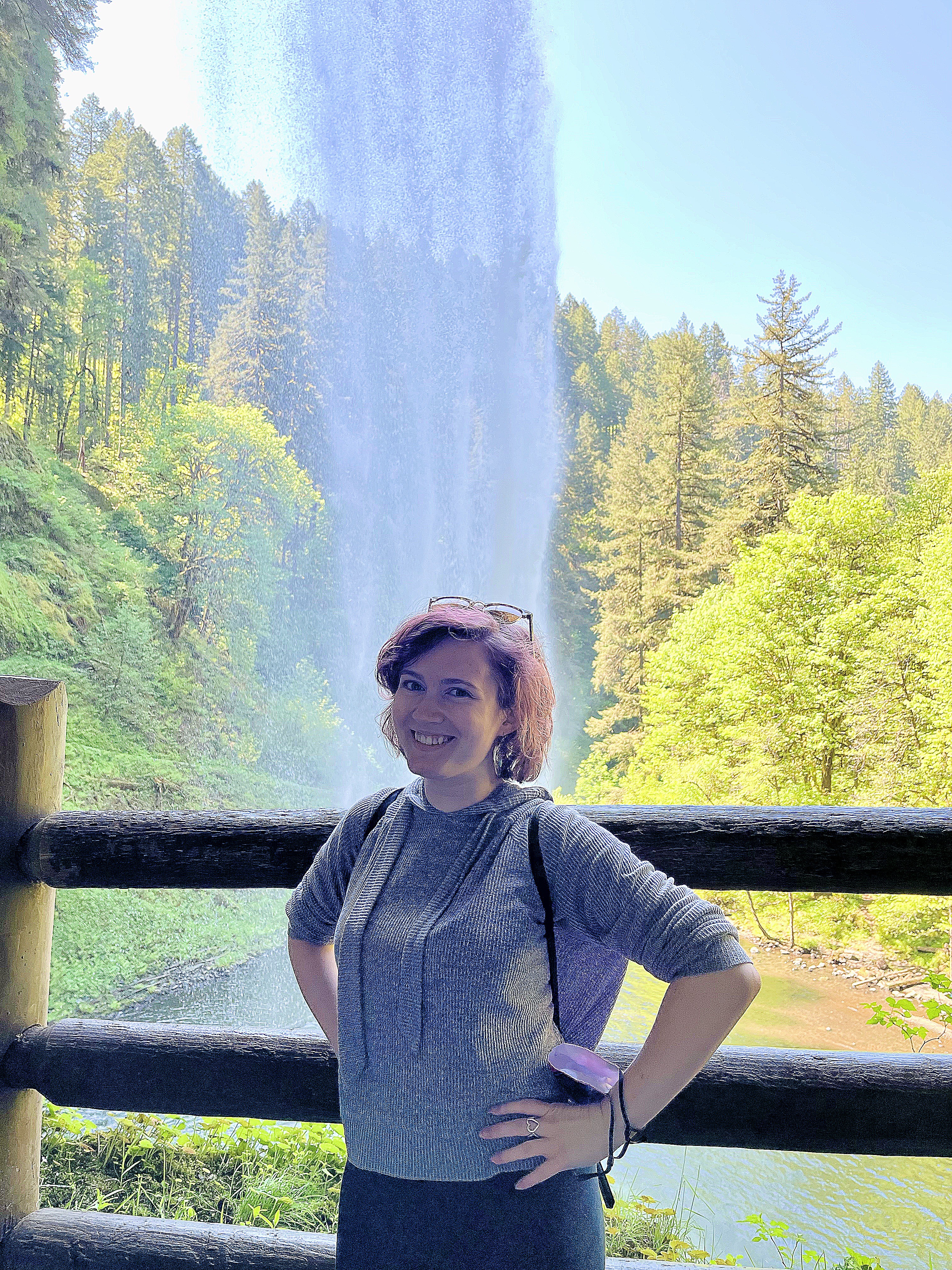 Sarah Grzyb (Sustainability Fellow) standing with hands on their hips and smiling in front of a waterfall.