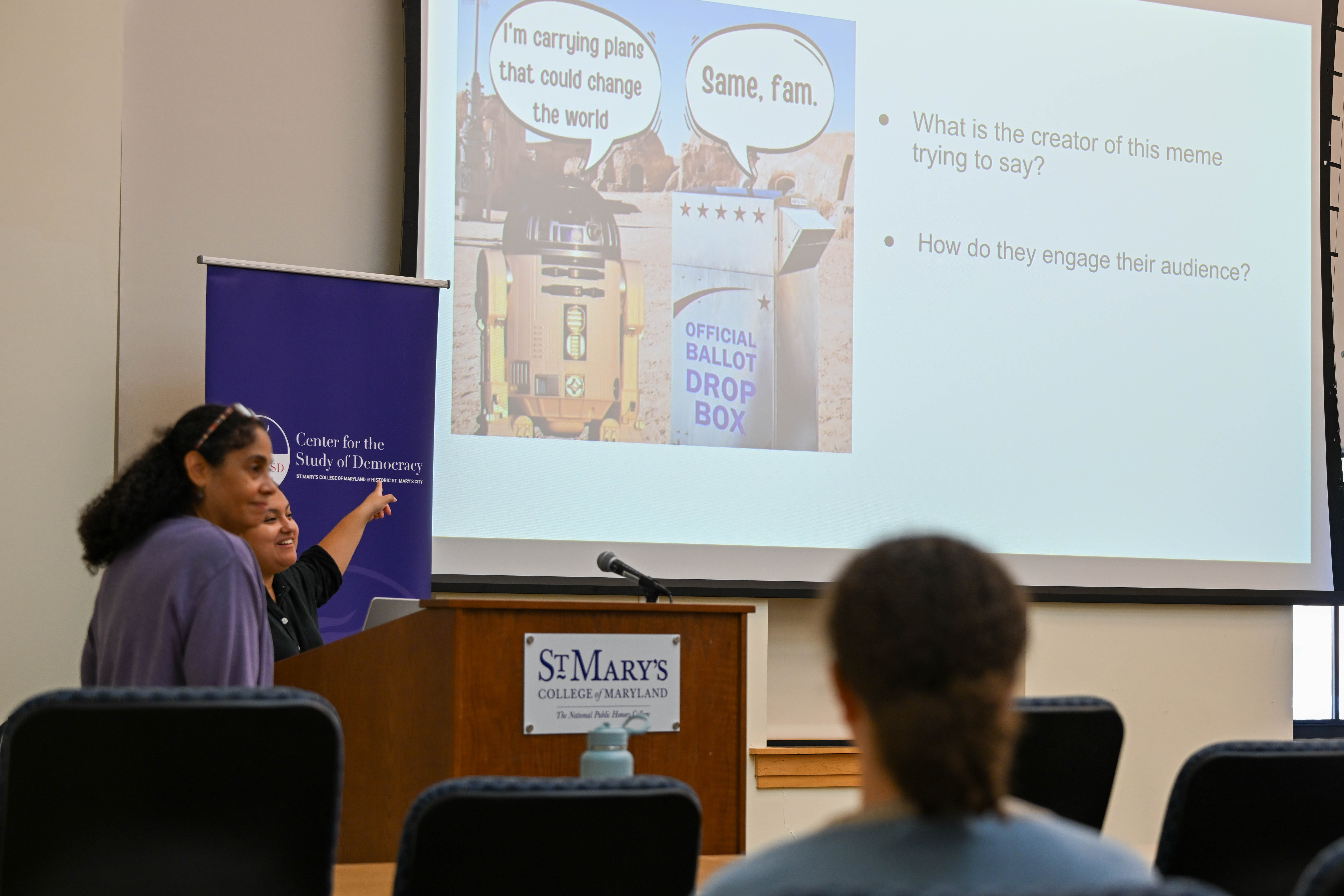 two people at a podium point to a meme on a big screen as the audience looks on. 