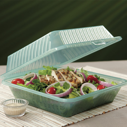 A half-opened, green reusable to-go box sits atop a white placemat on a gray table. The box contains a salad mixture and a small, plastic cup of salad dressing sits off to the side. The backdrop is dark green.