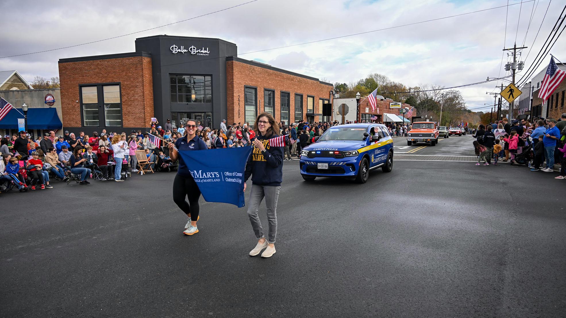 Two women carrying SMCM banner march in parade in front of SMCM Public Safety cruiser