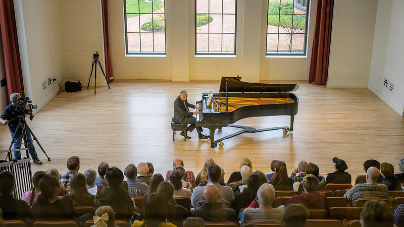 Man seated at piano before crowd in recital hall - video camera filming to the left