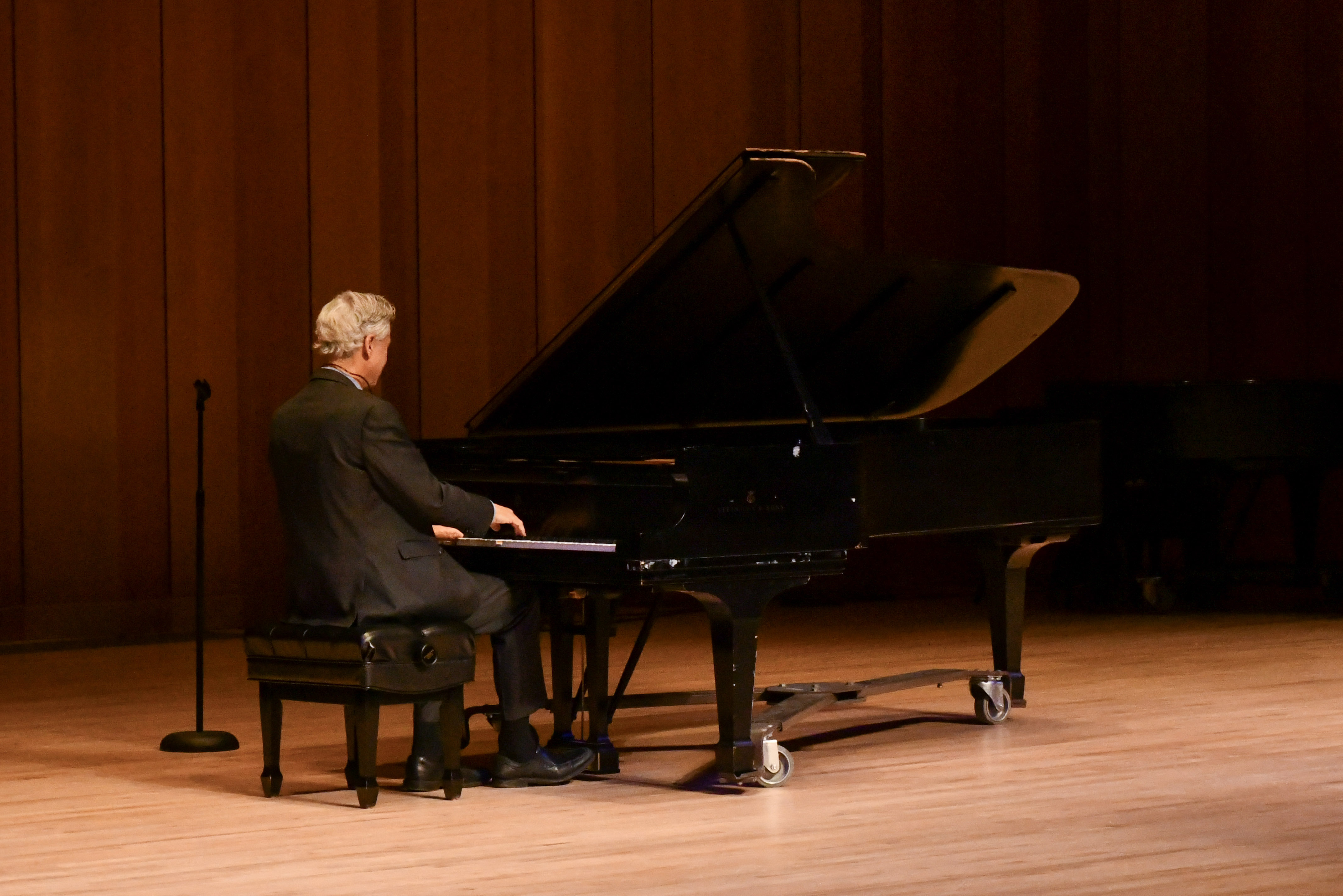 An adult with gray hair, dressed in a dark suit, is playing a grand piano on a wooden stage. A microphone stand is positioned to the left of the person, and the background features wooden paneling.