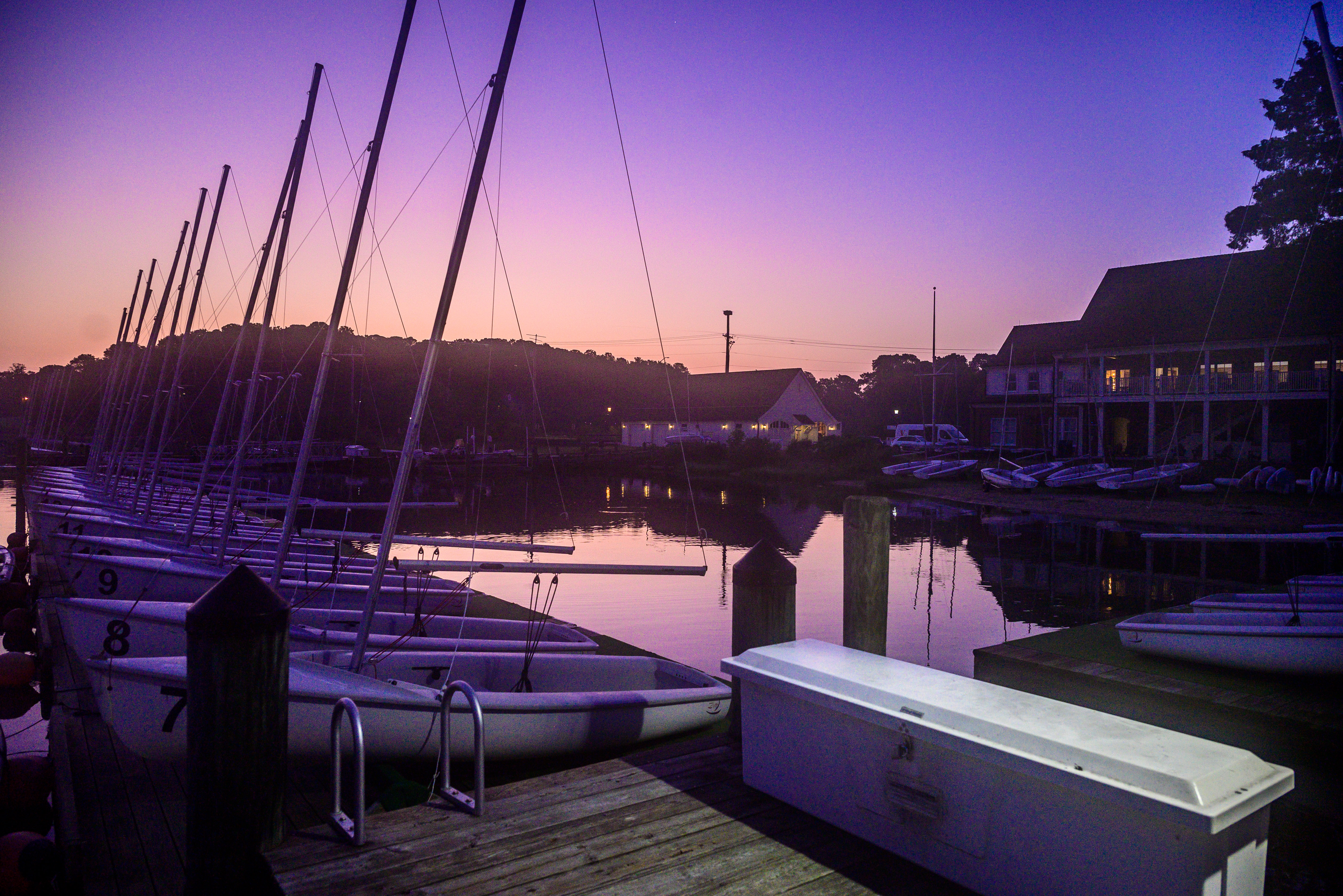 A serene marina at twilight with sailboats docked on the calm water. The sky transitions from orange to purple, reflecting on the water. Buildings and trees are silhouetted in the background. A white bench is on the wooden dock.