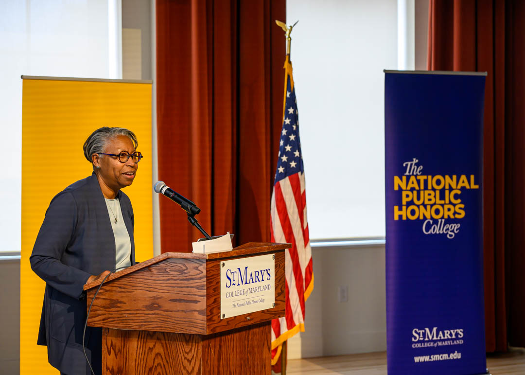 A person speaks at a podium labeled "St. Mary's College of Maryland," with an American flag and banners for the National Public Honors College and St. Mary's College in the background.