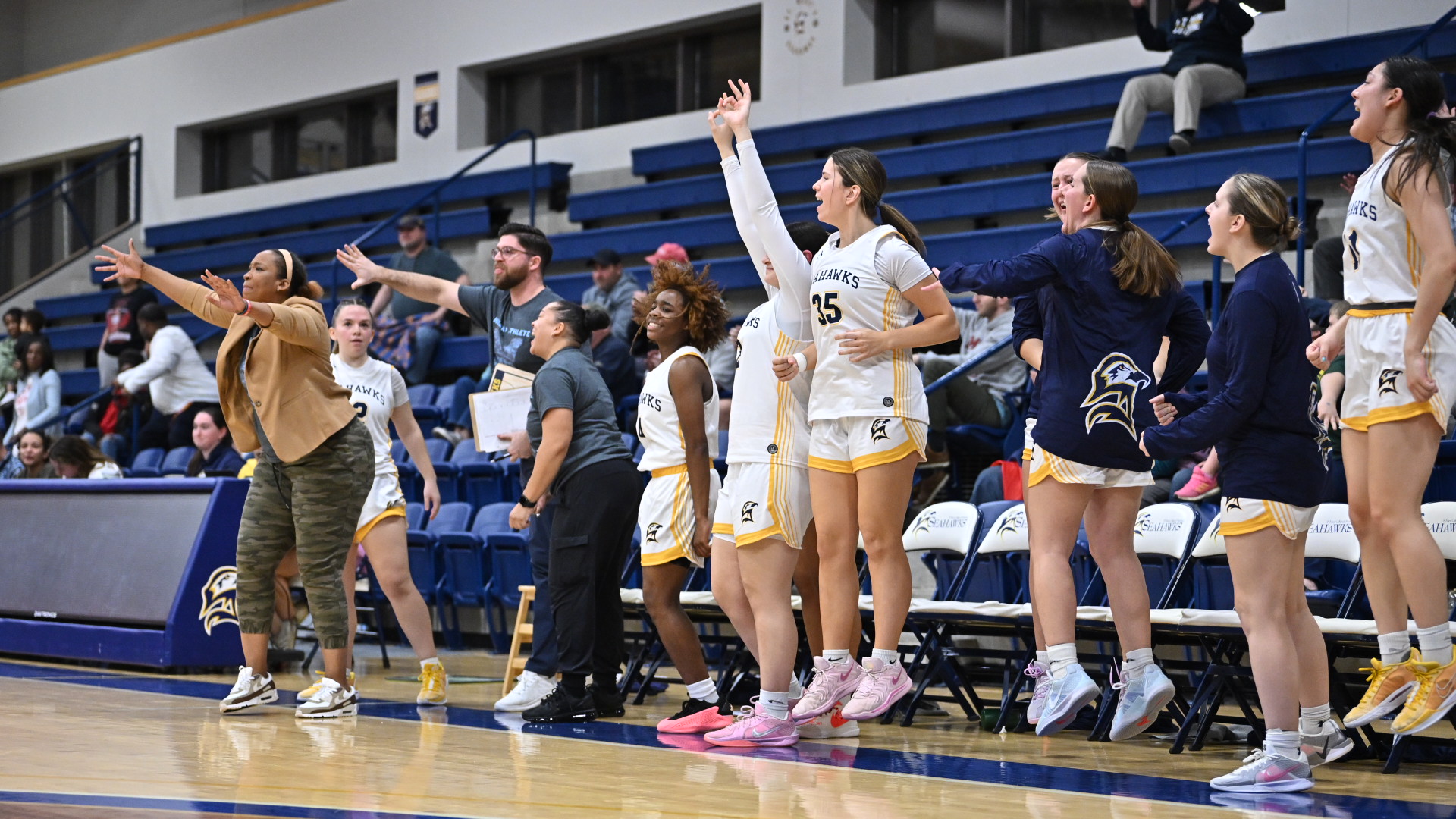 A women's basketball team celebrates on the sideline, some jumping with excitement. Coaches and players extend their arms enthusiastically. They wear white and yellow uniforms, with some in tracksuits. The bleachers are partially filled with spectators.