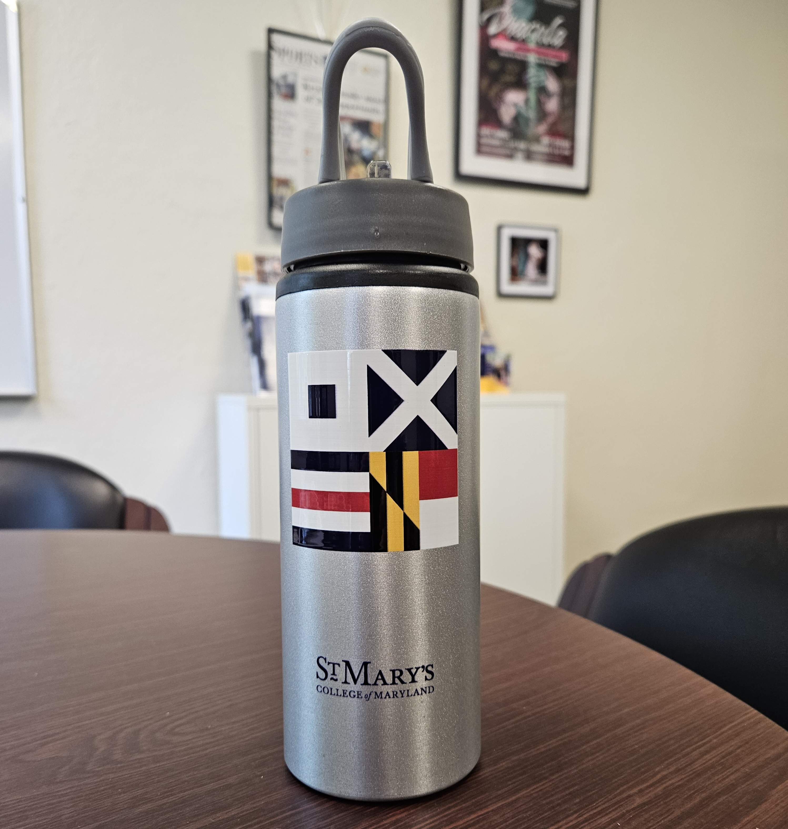 Silver water bottle with a dark lid on a round table. The bottle features colorful flag patterns and the text "St. Mary's College of Maryland." Blurred background includes framed posters and photos on a wall.