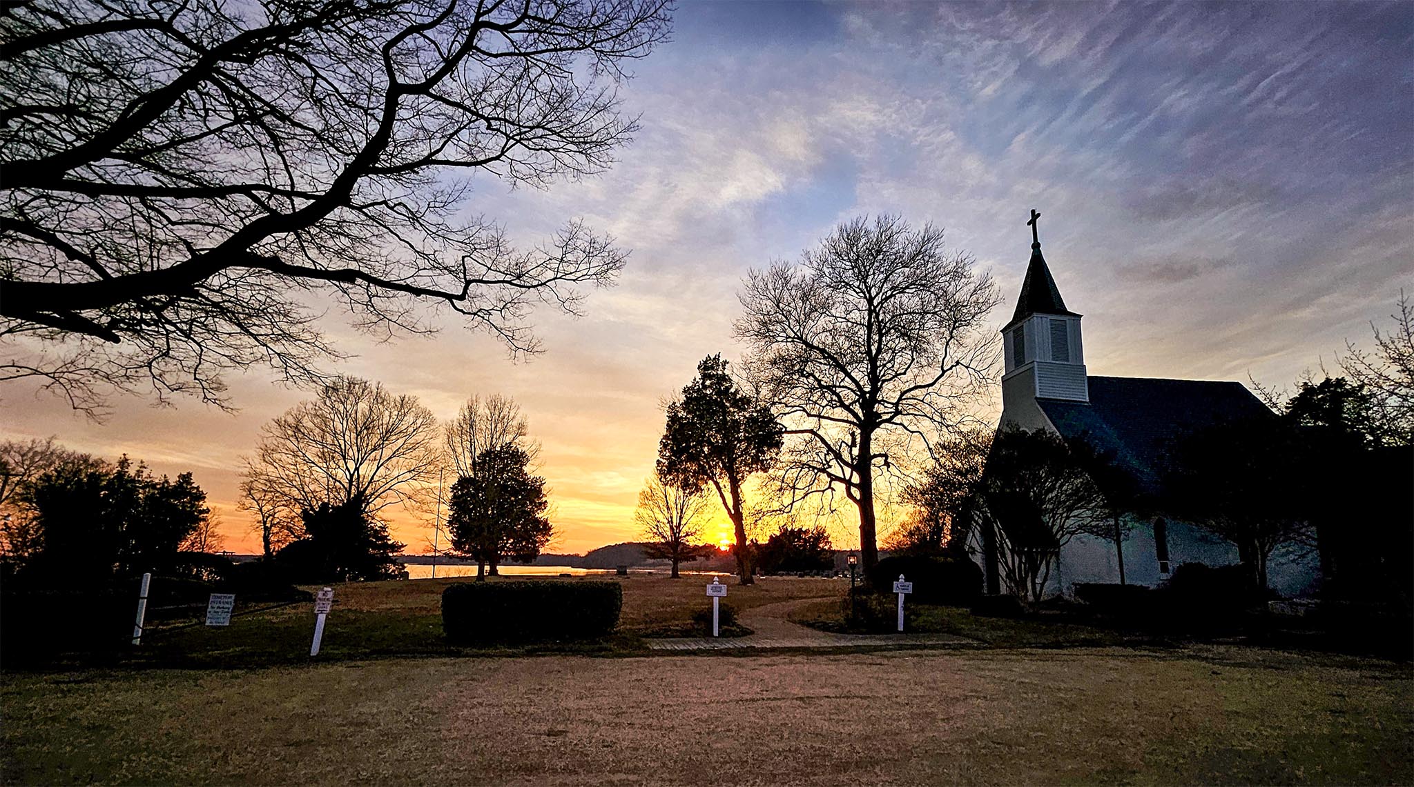 A serene scene of a church with a steeple silhouetted against a colorful sunset sky. Bare trees frame the foreground, and a body of water and hills are visible in the distance.