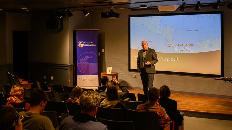 A person stands on a small stage giving a presentation to an audience. A projector screen shows a map including the Panama Canal Zone. A banner is visible on the left side of the stage. People are seated and facing the speaker.