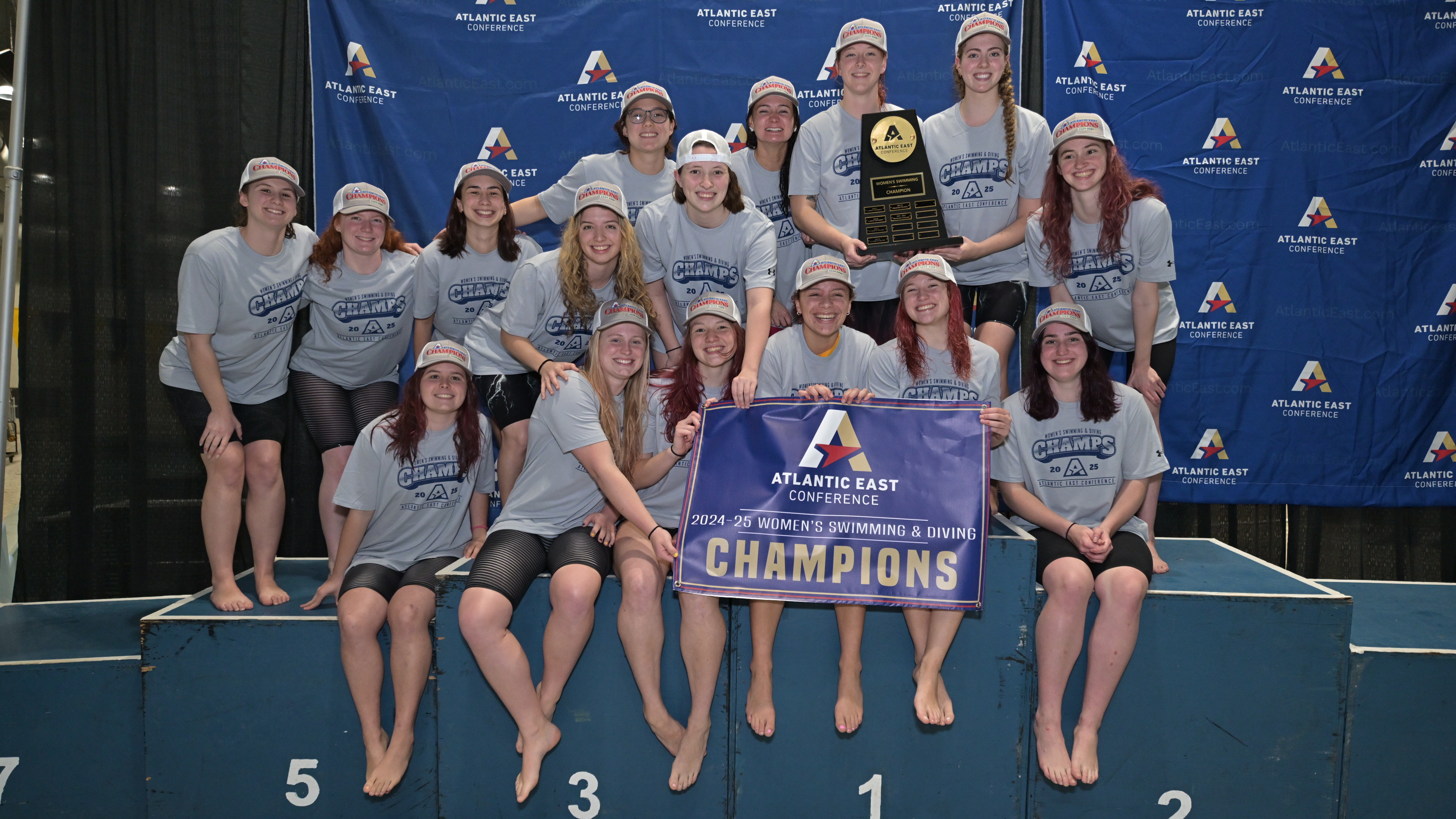 A women's swim team poses together wearing matching shirts and caps, holding a trophy and a banner that reads "Atlantic East Conference 2024-25 Women's Swimming & Diving Champions" against a branded backdrop.