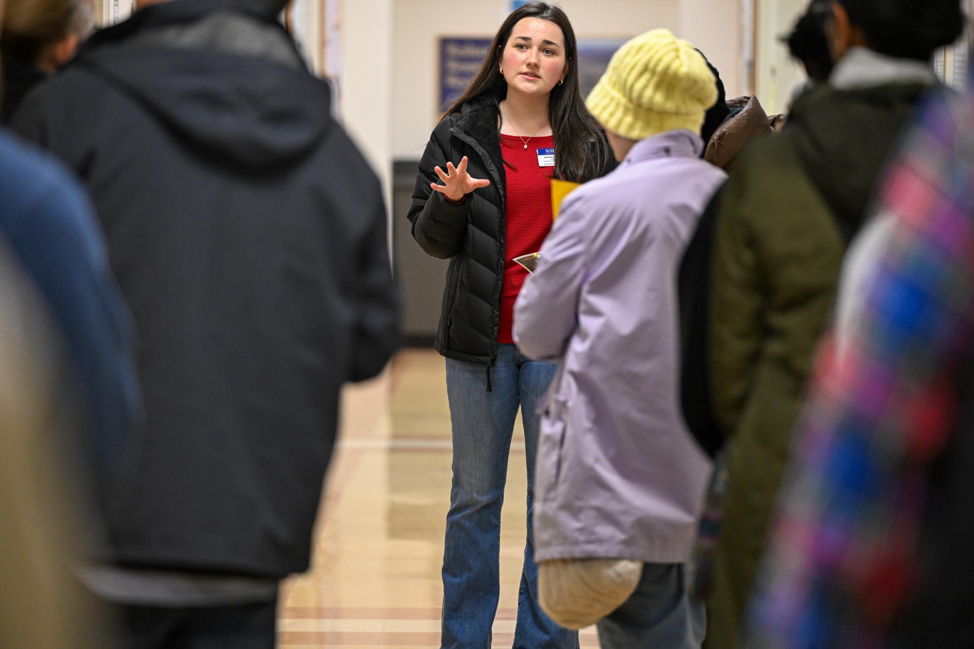 A woman stands in a hallway, gesturing with one hand as she speaks to a group of people. She has long dark hair, wears a red top and blue jeans, and has a name tag. The group is listening attentively, some wearing winter clothing.