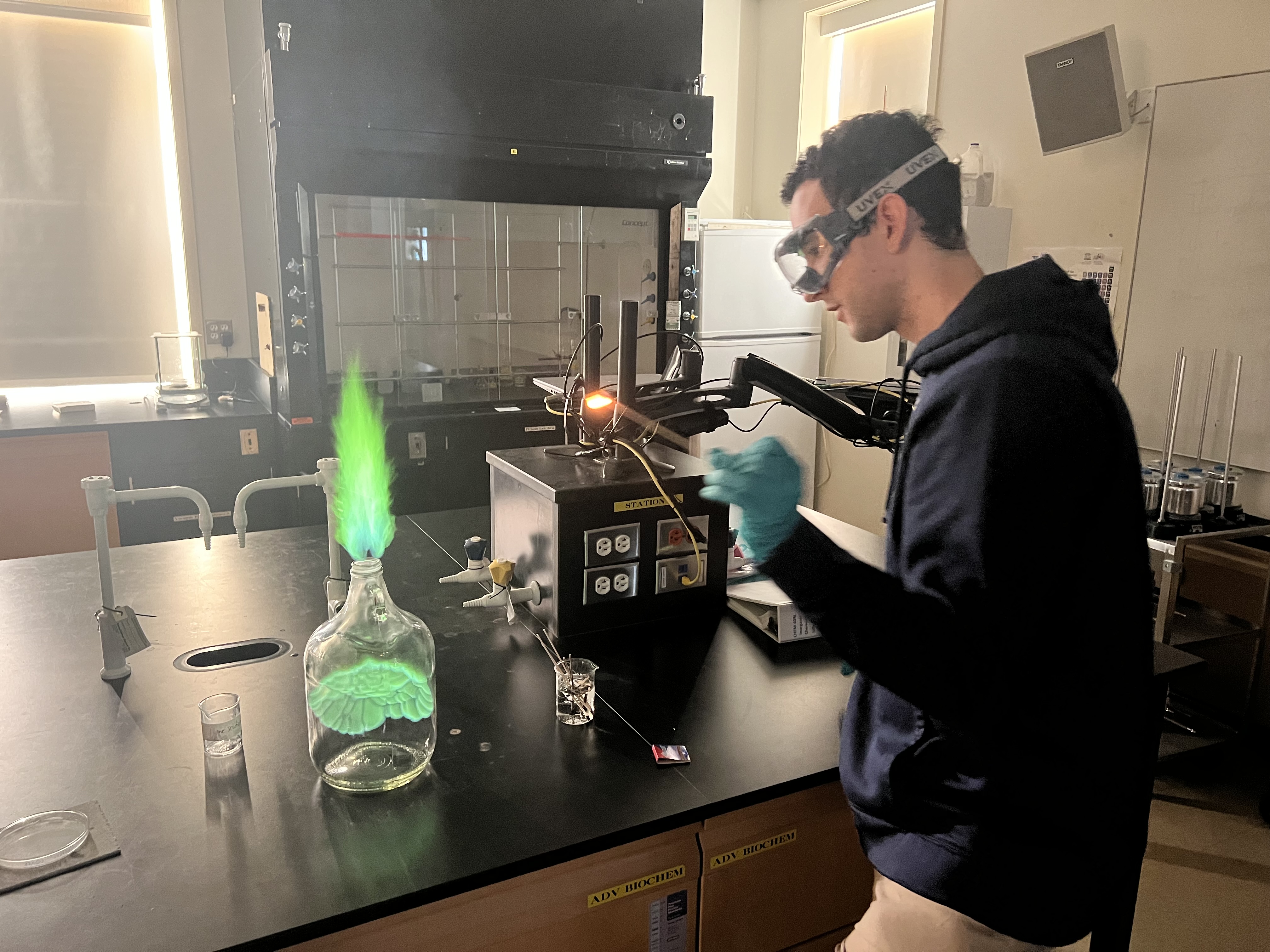 A person wearing safety goggles and gloves conducts an experiment in a lab. They are observing a bottle with a bright green flame emerging from it on a black table. Other lab equipment is visible in the background.