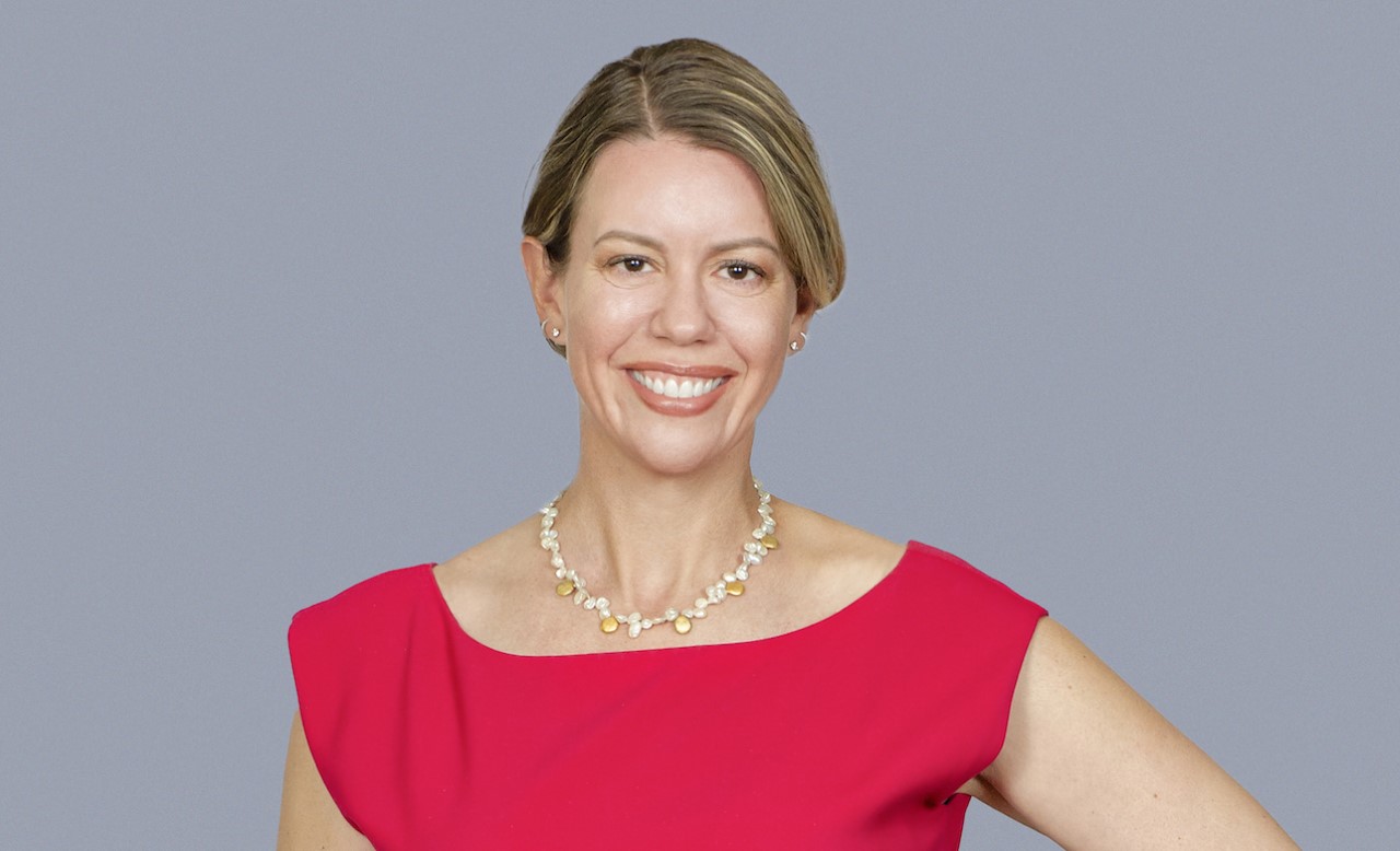 The speaker: Smiling woman with short blonde hair, wearing a bright red top and a pearl necklace, stands against a plain gray background.