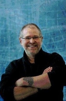 A person with glasses and a beard, wearing a dark shirt, smiles with arms crossed in front of a blue background.