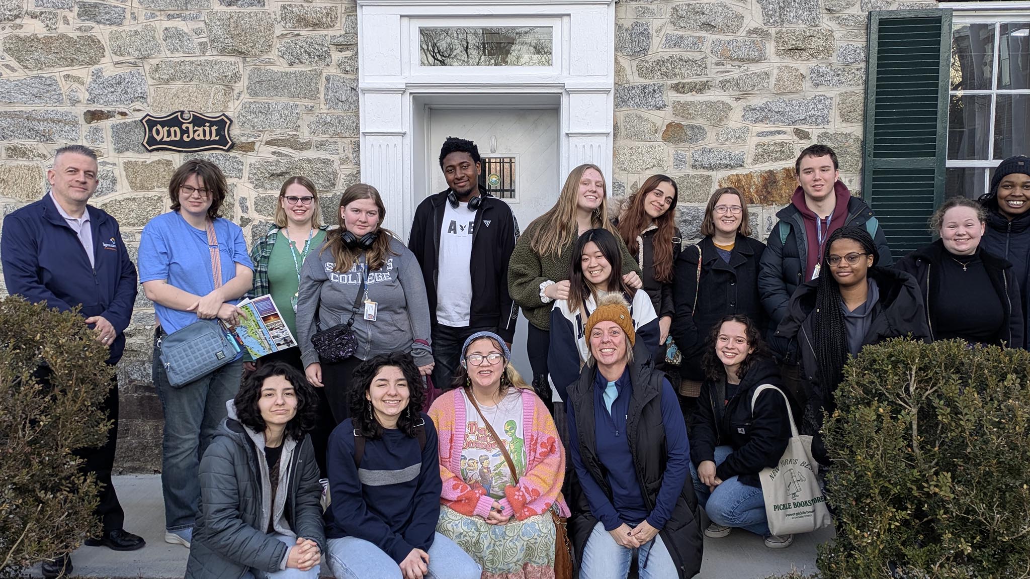 A group of people posing in front of a stone building with a white door and a sign that reads "Old Jail." Some are standing, and others are crouching. The group appears diverse in terms of gender and ethnicity. Vegetation is visible in the foreground.