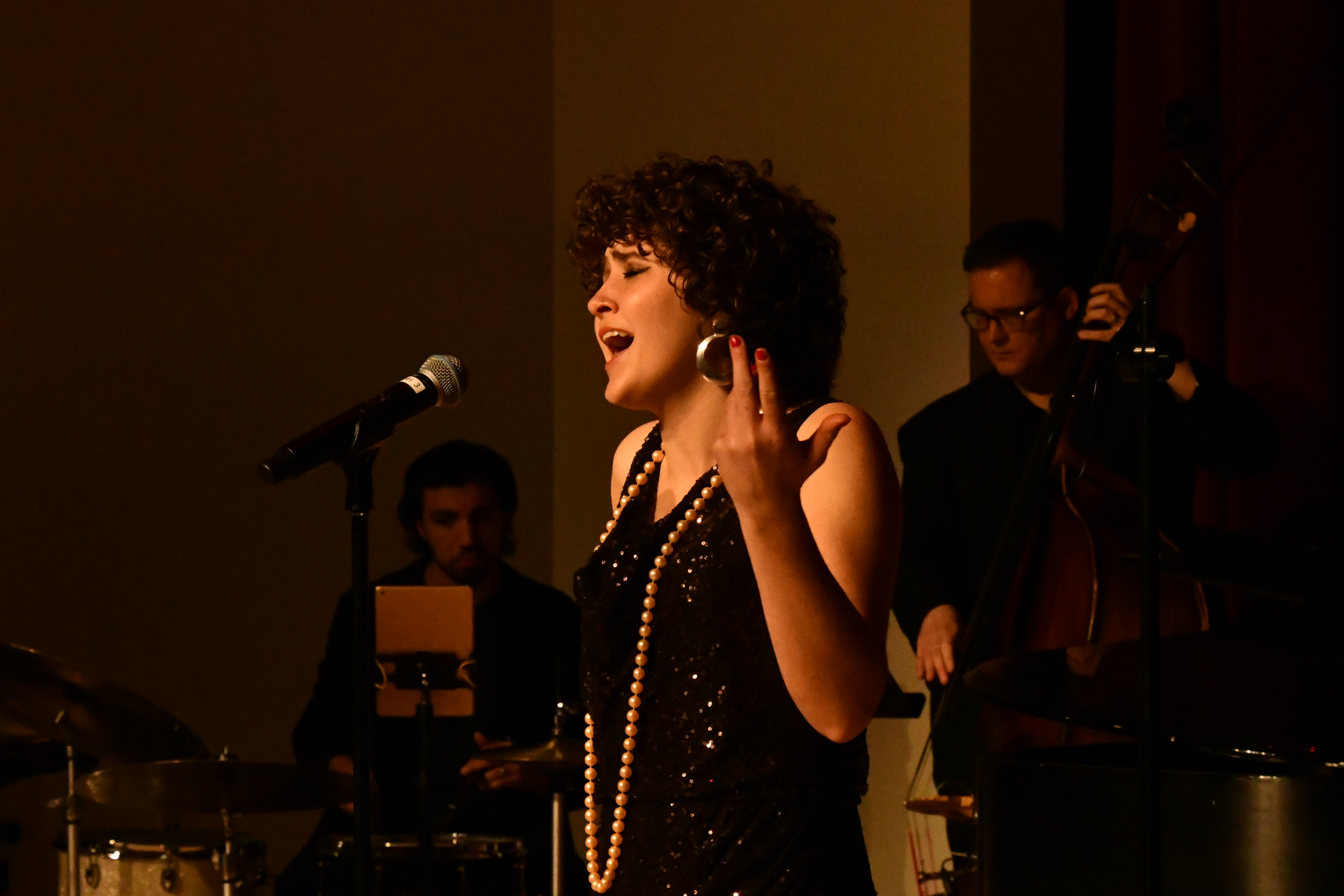 A woman with curly hair sings passionately into a microphone, wearing a black sequined dress and pearl necklace. A band plays in the background with a drummer and a double bassist. The scene is warmly lit, creating a cozy atmosphere.