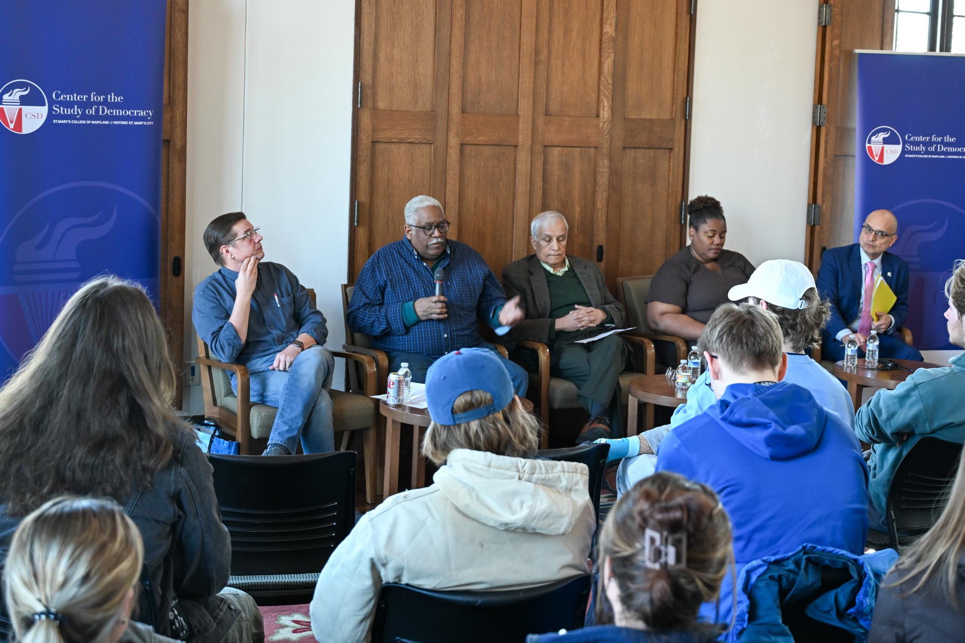 A panel of five people seated in chairs, engaged in a discussion. An audience listens attentively in a room with banners displaying "Center for the Study of Democracy." The panelists are diverse in appearance and age.