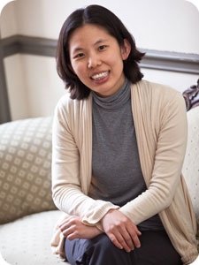 A person with shoulder-length dark hair (Belinda Kong) sits on a patterned couch, smiling. They are wearing a gray turtleneck and a beige cardigan, with their hands resting on their lap. The background features wainscoting and soft lighting.