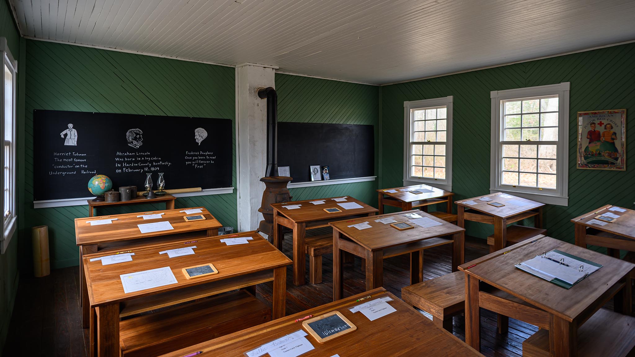 A vintage classroom with wooden desks, a blackboard displaying chalk drawings and text, old-fashioned stove, and educational posters. The room is lit by natural light from the large windows, showcasing a historical school setting.