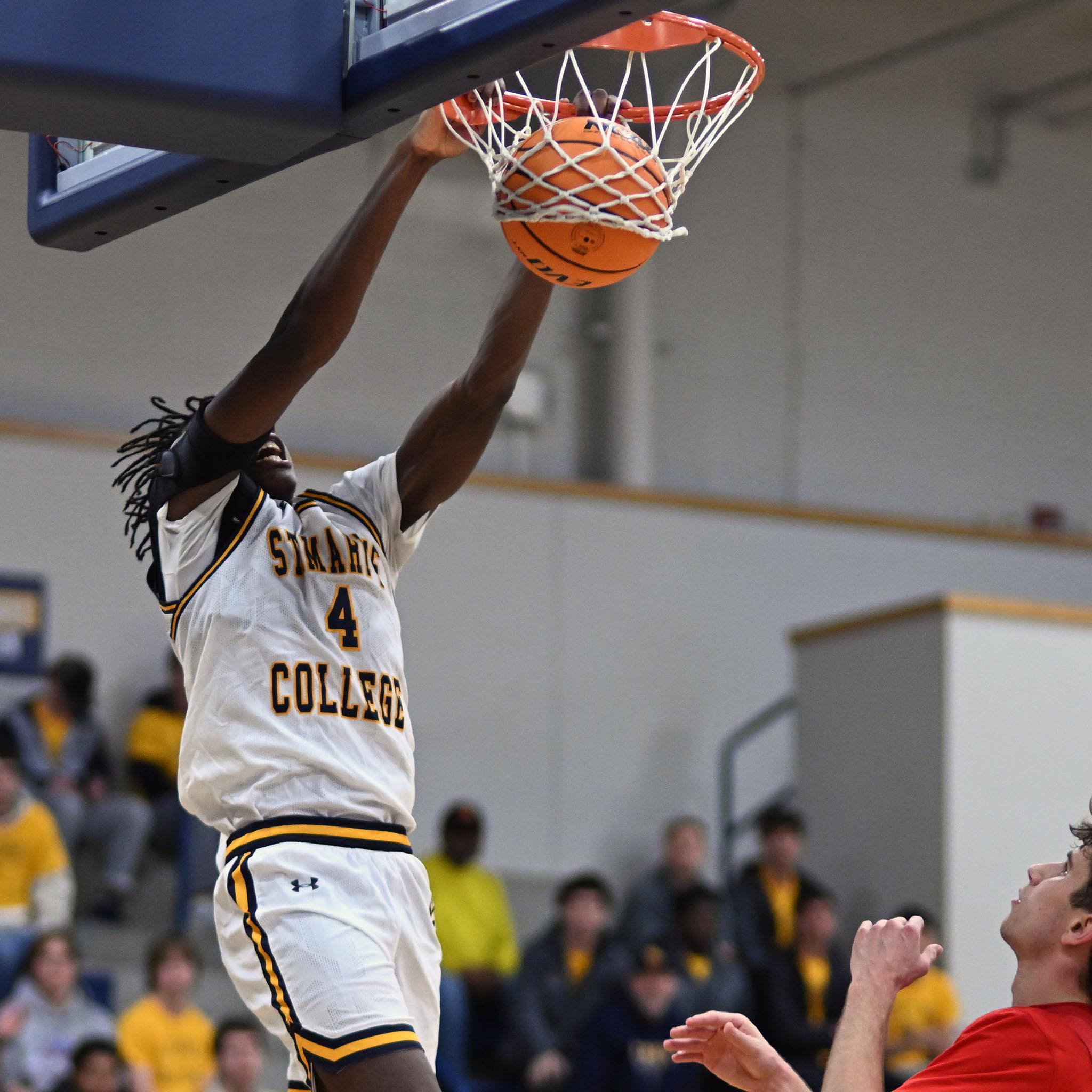 A basketball player wearing a white and gold "St. Mary's College" uniform, number 4, performs a slam dunk. Another player in a red jersey is visible below. Spectators in yellow shirts sit in the blurred background.