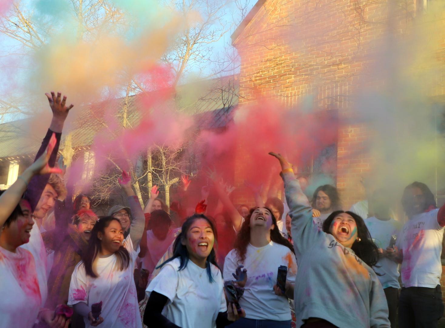 A group of people wearing white shirts joyfully celebrate by throwing colorful powder into the air. The vibrant colors contrast with the brick building and trees in the background. Smiles and laughter are seen as they enjoy the festive atmosphere.