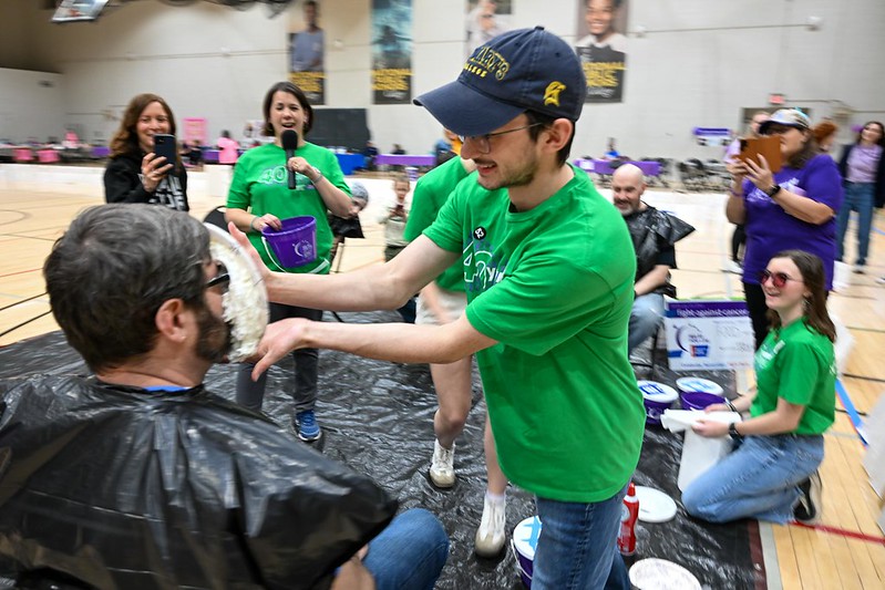 A young man in a green shirt playfully smashes a whipped cream pie into another man's face at an indoor event. The recipient wears glasses and a black plastic cover. Nearby, people watch and laugh, wearing casual clothing.