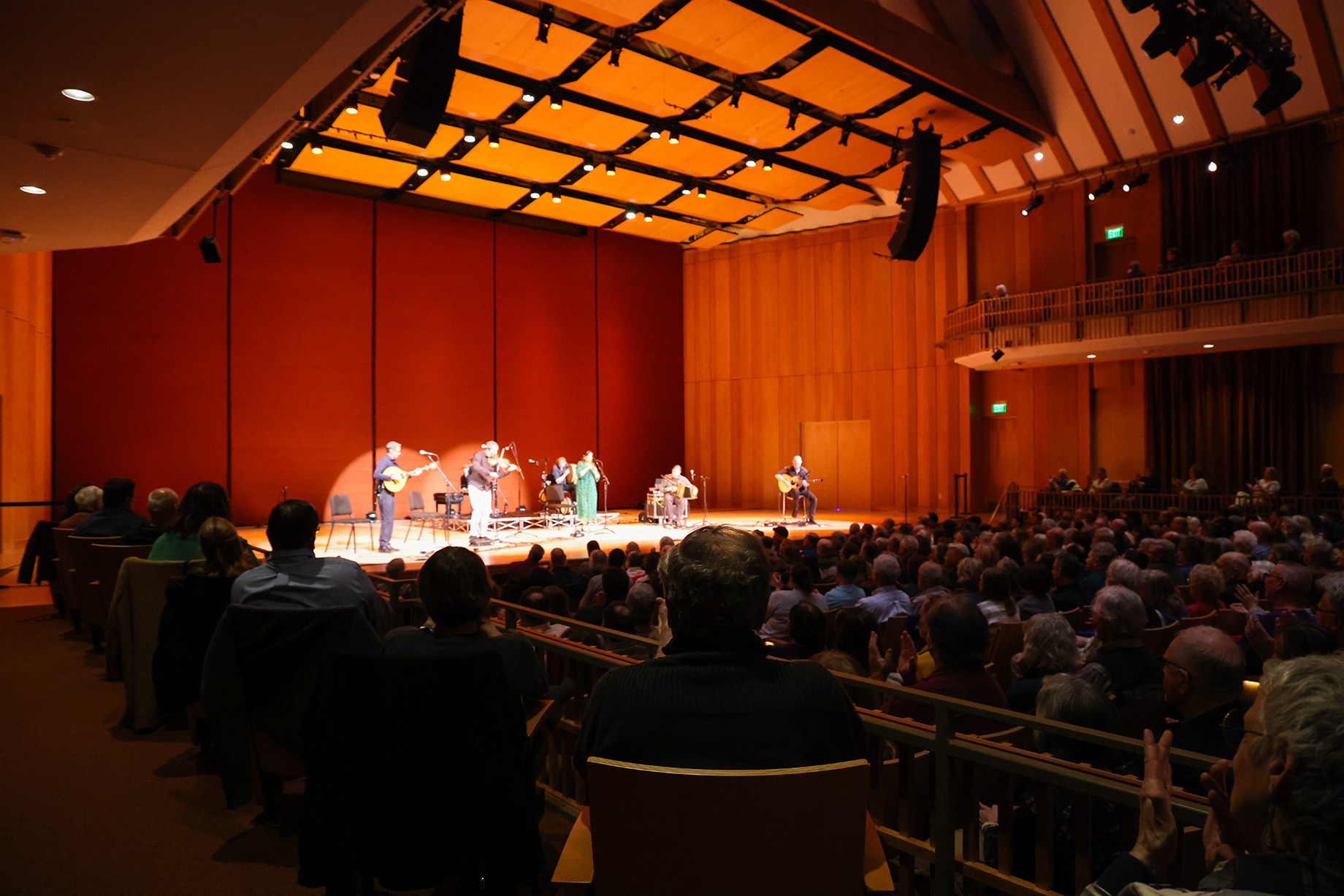 A musical performance is taking place on a well-lit stage in a large auditorium. Musicians play various instruments as a full audience watches. The walls and ceiling are wooden, enhancing the room's acoustics.