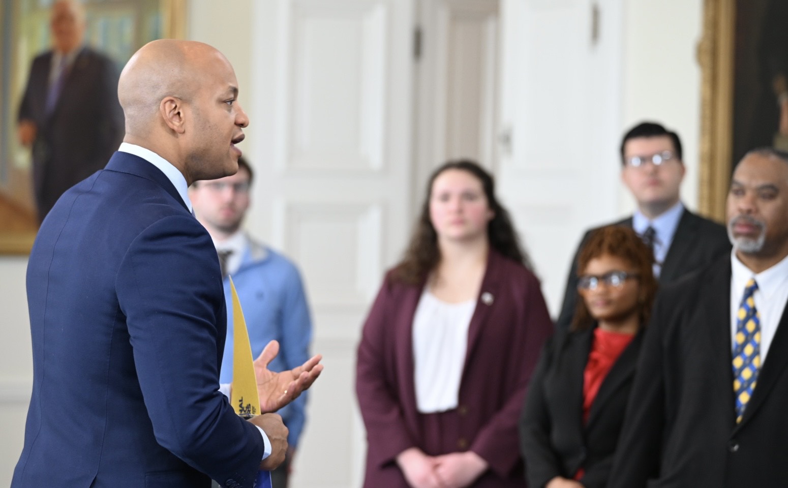 A man in a blue suit speaks to a group of people indoors. The group includes individuals in formal attire, standing attentively. A portrait hangs on the wall in the background, and there's natural lighting in the room.