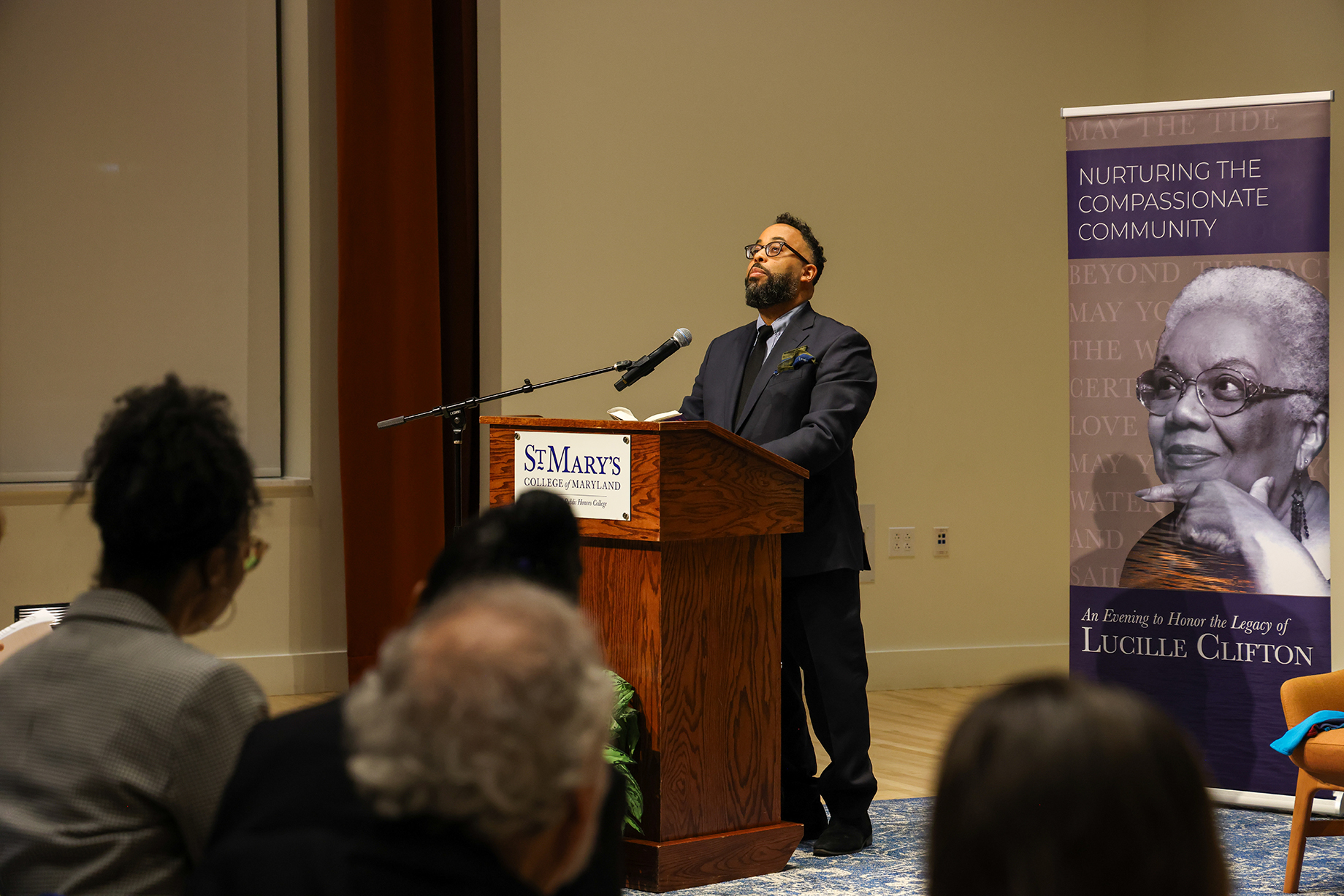 A man stands at a podium giving a speech in a room with a banner featuring Lucille Clifton that reads "Nurturing the Compassionate Community." Audience members are seated in the foreground.