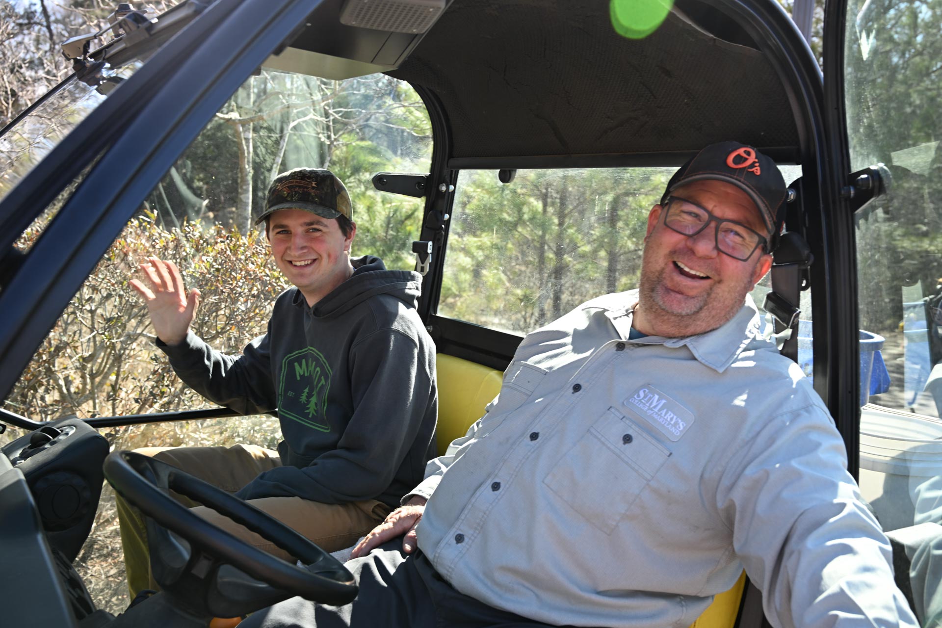 Two men sit in a vehicle, facing the camera. The man on the left waves and wears a cap and hoodie. The man on the right, also in a cap, smiles broadly. They appear relaxed and outdoors, surrounded by trees and bushes.