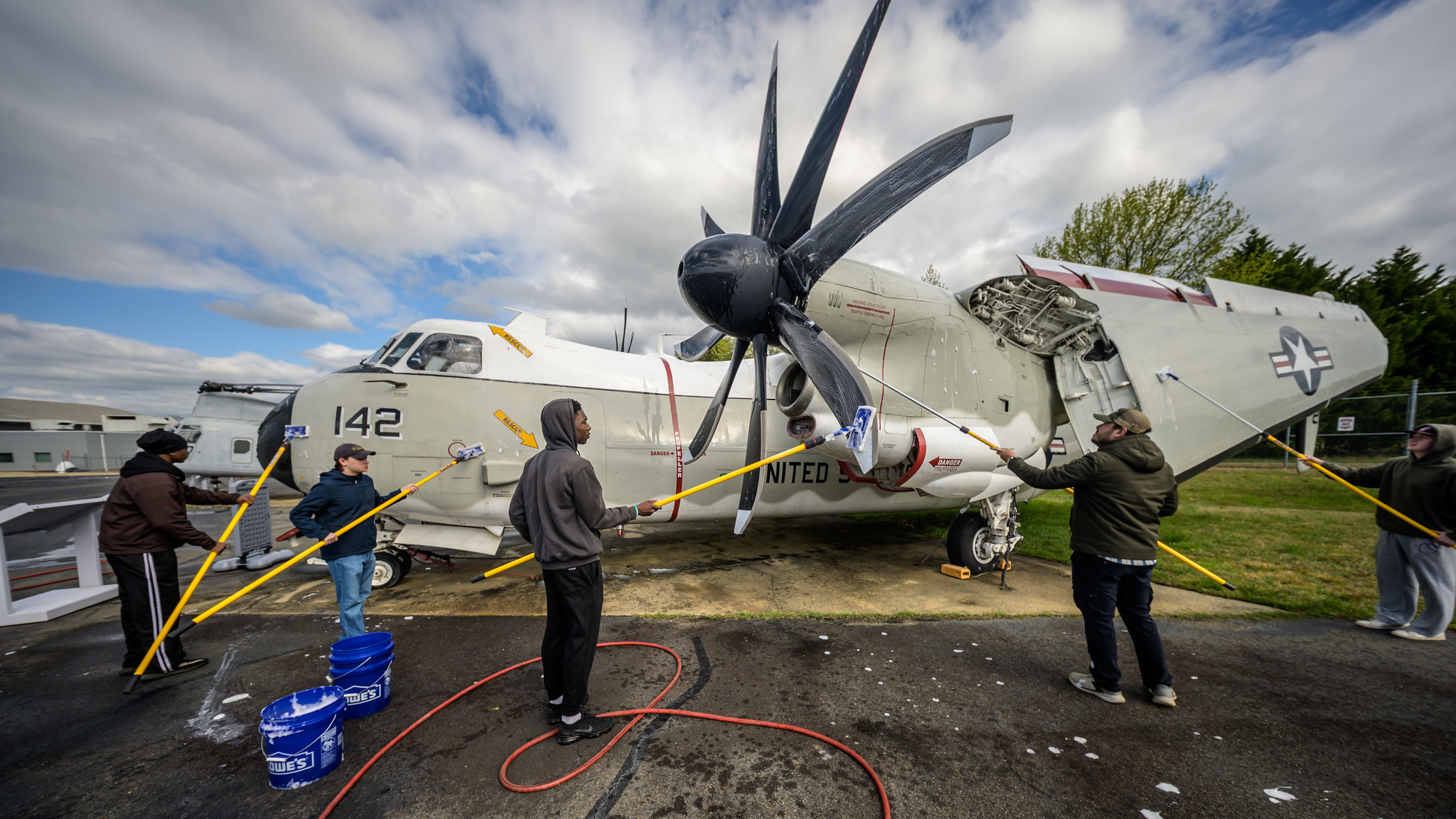 Several people wash a large military aircraft with mops and brushes outdoors, with cleaning supplies and buckets visible around them.