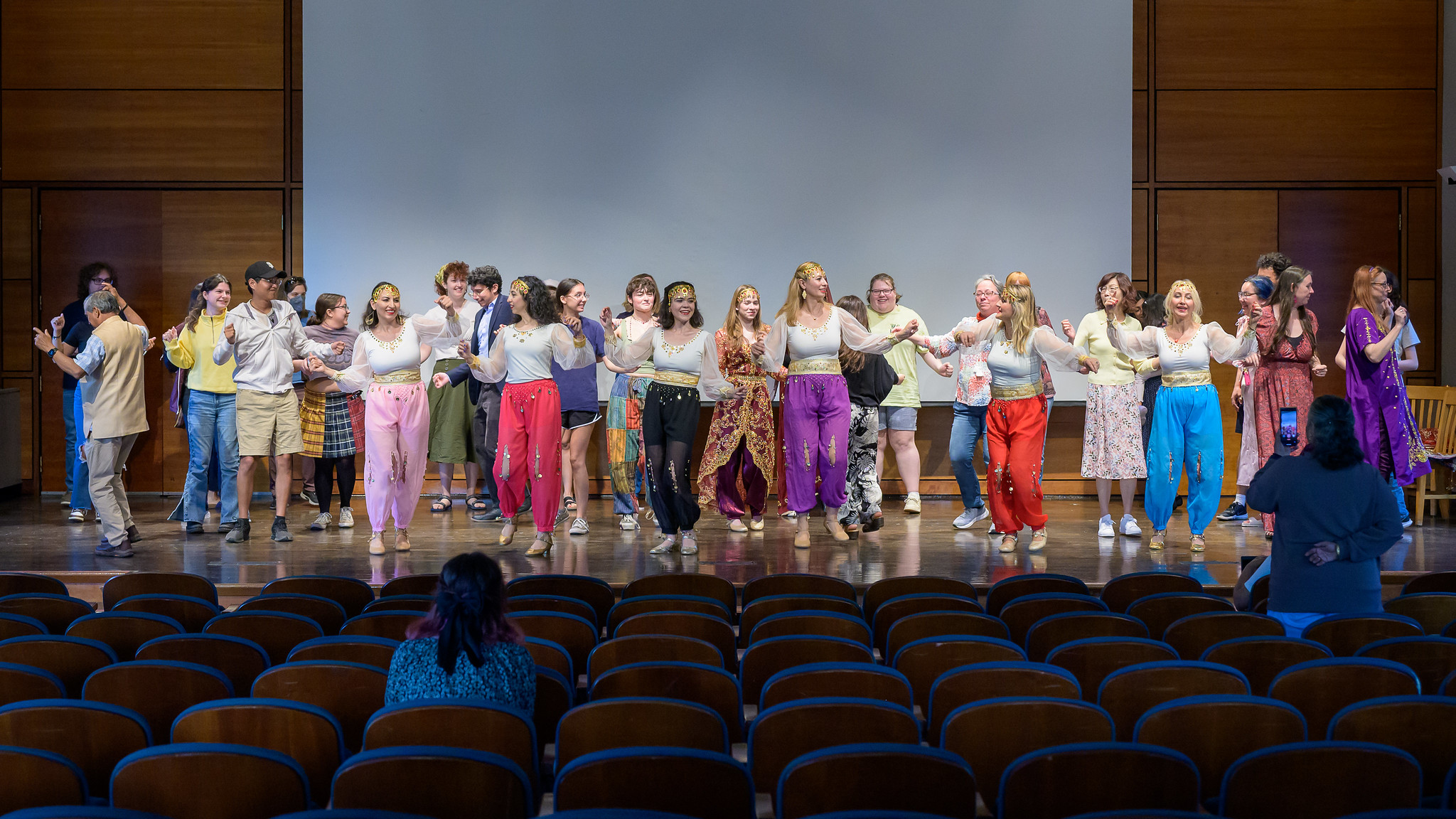 A group of people perform a group dance on a stage in front of an audience in a wooden auditorium.
