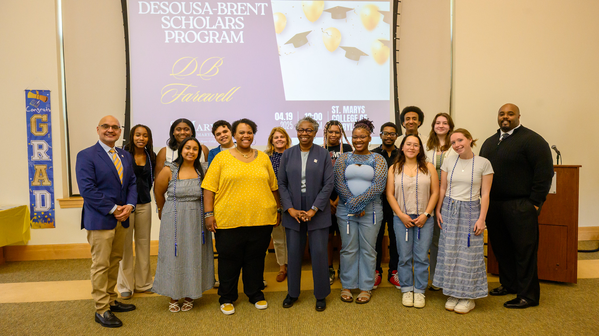A group of students and staff pose for a photo in front of a screen displaying "Desousa-Brent Scholars Program" during an indoor event.