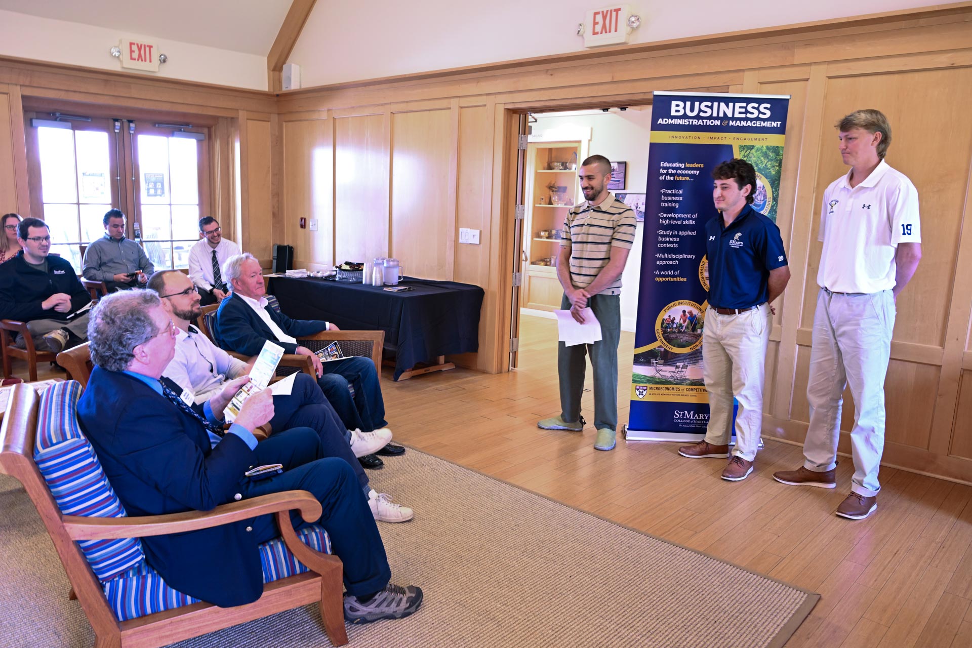 Three young men stand and present to a seated group in a wood-paneled room, with a business program banner visible in the background.