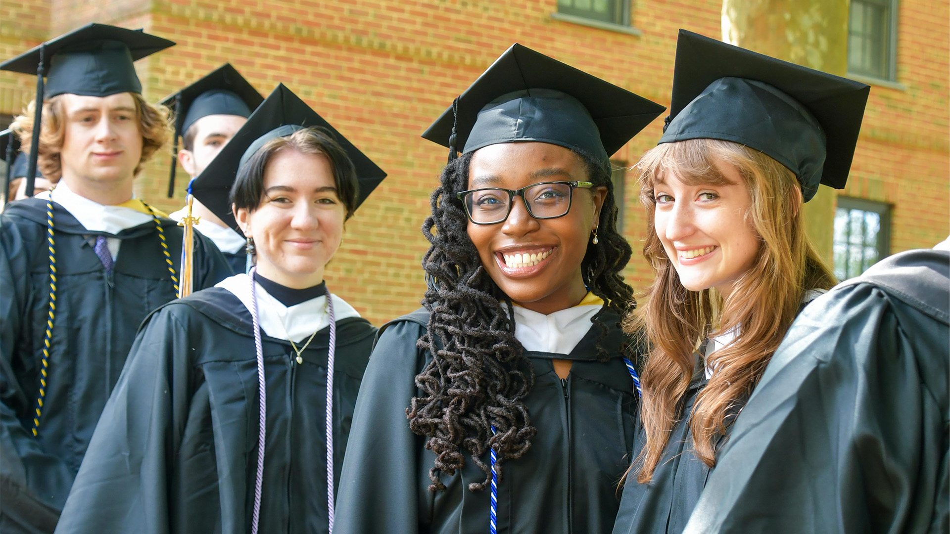 A group of students wearing graduation caps and gowns stand in a line outdoors, smiling at the camera.