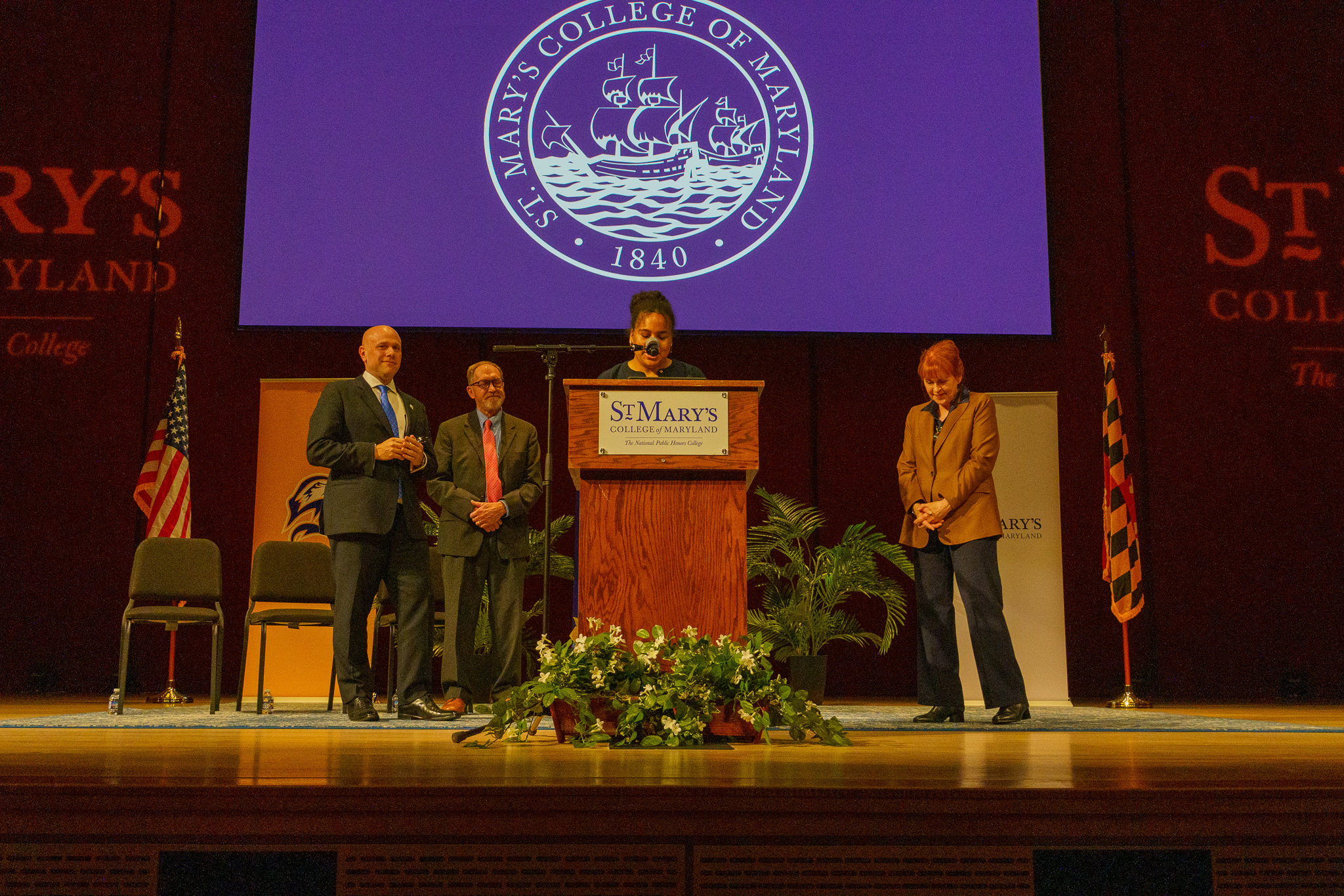 Four people stand on stage at a St. Mary’s College of Maryland event, with a speaker at a podium and the college seal projected in the background.